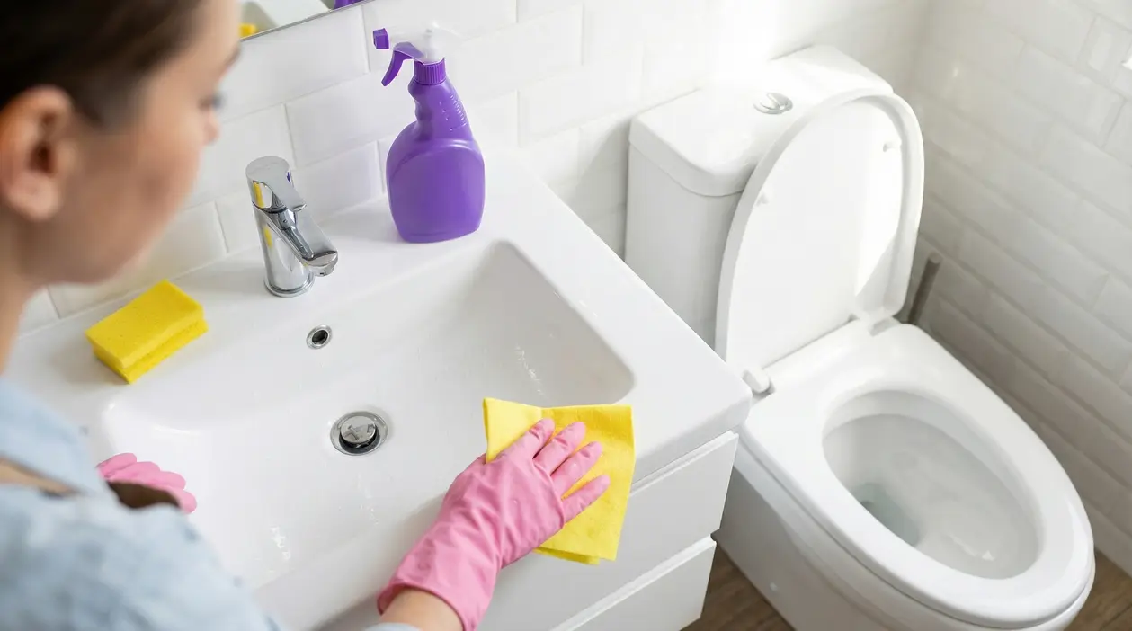 Persona che pulisce il lavandino del bagno con un panno giallo e guanti rosa