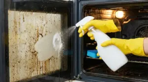 Persona con guanti gialli spruzza detergente sulla porta incrostata di un forno aperto